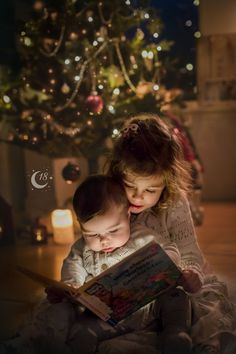 two young children reading a book in front of a christmas tree with lights on it