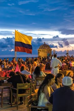 many people are sitting at tables on the beach and one person is holding a flag