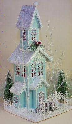 a blue and white christmas house with snow on the roof, trees and fence around it