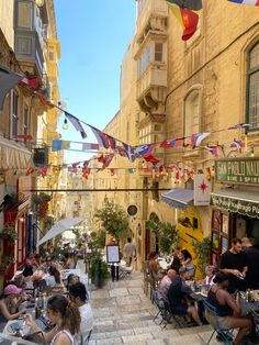 people sitting at tables in an alleyway with flags hanging from the buildings behind them