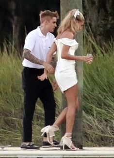 a man and woman walking next to each other near the water in white dress with high heels