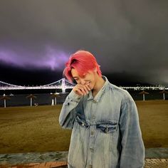 a woman with pink hair standing in front of a body of water under a cloudy sky