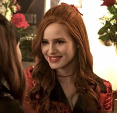 a woman with long red hair is talking to another woman in a room full of flowers