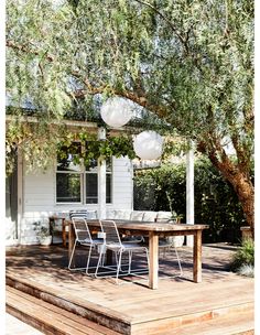 a table and chairs on a wooden deck under a tree in front of a white house