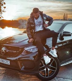 a man sitting on the hood of a black mercedes c - class parked in front of a lake