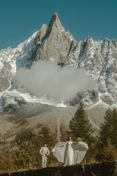 a bride and groom standing in front of a mountain with their wedding dress blowing in the wind