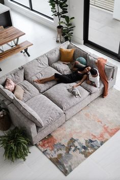 two people sitting on a large gray couch in the middle of a living room with lots of windows