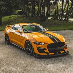 an orange sports car with black stripes parked in a parking lot next to some trees