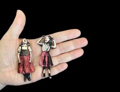 two dolls are sitting on the palm of a person's hand, one is wearing a red dress