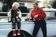 two people sitting on the hood of an old car, one holding a cup and talking