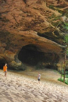 two people are walking on the beach near a cave