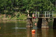 two boys are jumping into the water from a bridge over a body of water with trees in the background