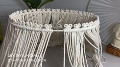 a white macrame lamp shade with chains hanging from it's sides on a table next to a busturine