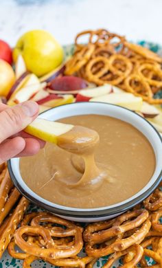 a person dipping caramel into a bowl of apple cider sauce with pretzels and apples in the background