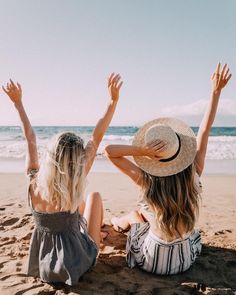 two women sitting on the beach with their arms in the air