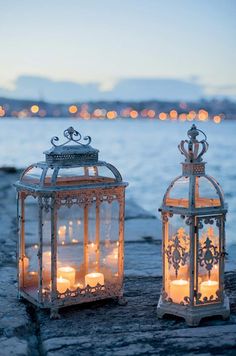 two lanterns with lit candles in them sitting next to each other on the ground near water