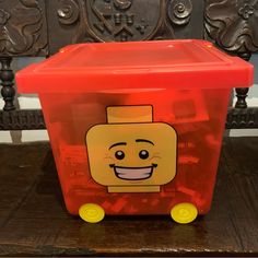 a plastic toy box with a lego face on the lid and wheels, sitting on a wooden table