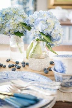 blue and white flowers in glass vases on a table set with plates, silverware and utensils