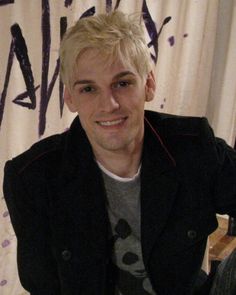 a young man with blonde hair is smiling for the camera while sitting in front of a curtain