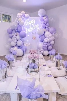 a table set up for a birthday party with purple and white balloons on the wall