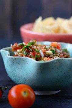 two bowls filled with food sitting next to some tomatoes and chips on a blue table