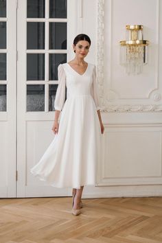 a woman wearing a white dress standing in front of a wall with glass doors and a chandelier