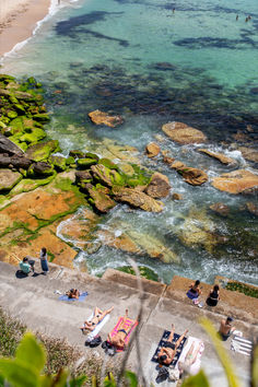 people are sitting on the rocks near the water and in the sand at the beach