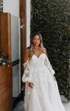 a woman wearing a white wedding dress standing in front of a wall with greenery