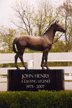 a statue of a horse stands in front of a white fence with trees behind it