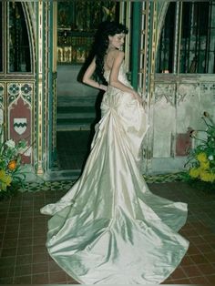 a woman in a long white dress is standing near a wall with flowers on it