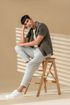 a young man sitting on top of a wooden stool next to a white wall and holding his hand under his chin