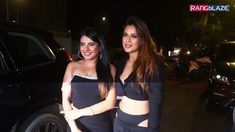 two beautiful women standing next to each other in front of a black car at night