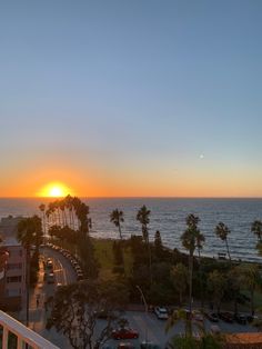 the sun is setting over the ocean with cars parked on the street and palm trees