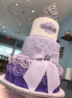 a three tiered cake decorated with purple and white roses, bows and a tiara