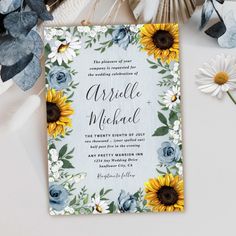 a wedding card with sunflowers and blue flowers on it next to some feathers