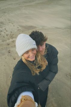 two people are hugging on the beach with their arms around each other and one is wearing a beanie