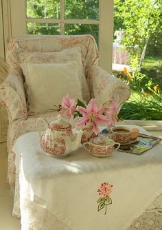 a table with tea cups and flowers on it in front of a chair near a window