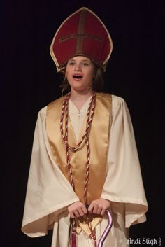 a young boy dressed in a priest outfit and holding a cross with his hands, standing on stage