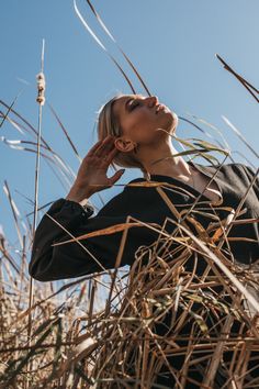 a woman standing in tall grass with her hands to her ear and looking up into the sky