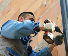 a man in blue jacket holding a drill and hammer next to a pole with brick wall behind him