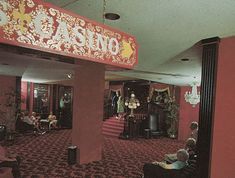 people are sitting and standing in a casino lobby with red carpeted walls, black chairs, and chandeliers