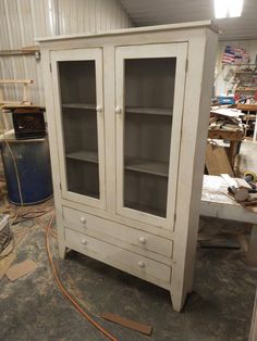 an old white china cabinet with glass doors in a workshop area, surrounded by tools and materials