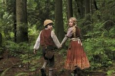 two people dressed in period clothing walking through the woods holding hands and looking at each other