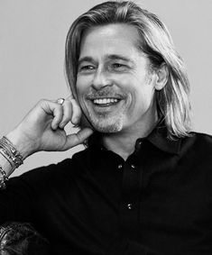 a black and white photo of a man with long hair smiling at the camera while sitting down