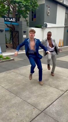 two men are dancing on the sidewalk in front of a building and one is wearing a blue suit