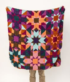 a person holding up a colorful quilt on top of a white wall with brown boots