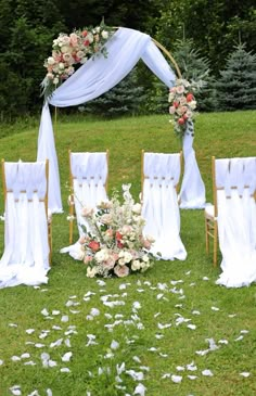 an outdoor ceremony setup with white drapes and flowers on the grass, surrounded by wooden chairs