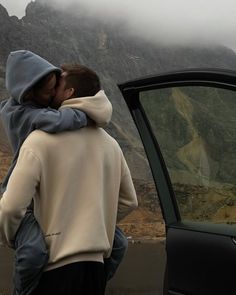 two people standing next to each other near a car with mountains in the back ground