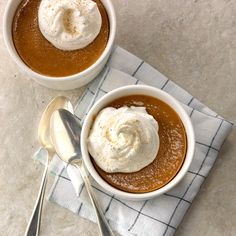 two white bowls filled with caramel pudding on top of a table next to silver spoons