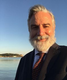 an older man wearing a suit and tie standing in front of the water with his arms crossed